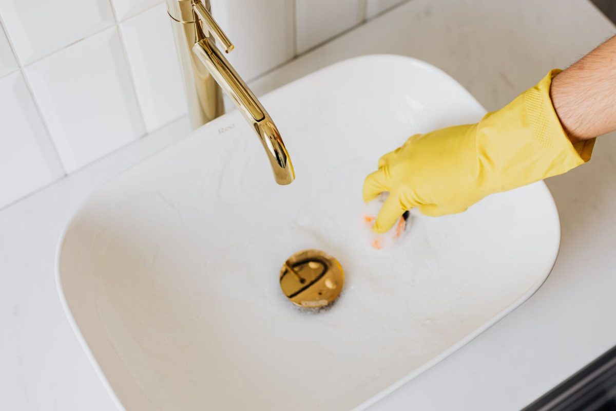 Gloved hand cleaning a white bathroom sink with a gold faucet in view