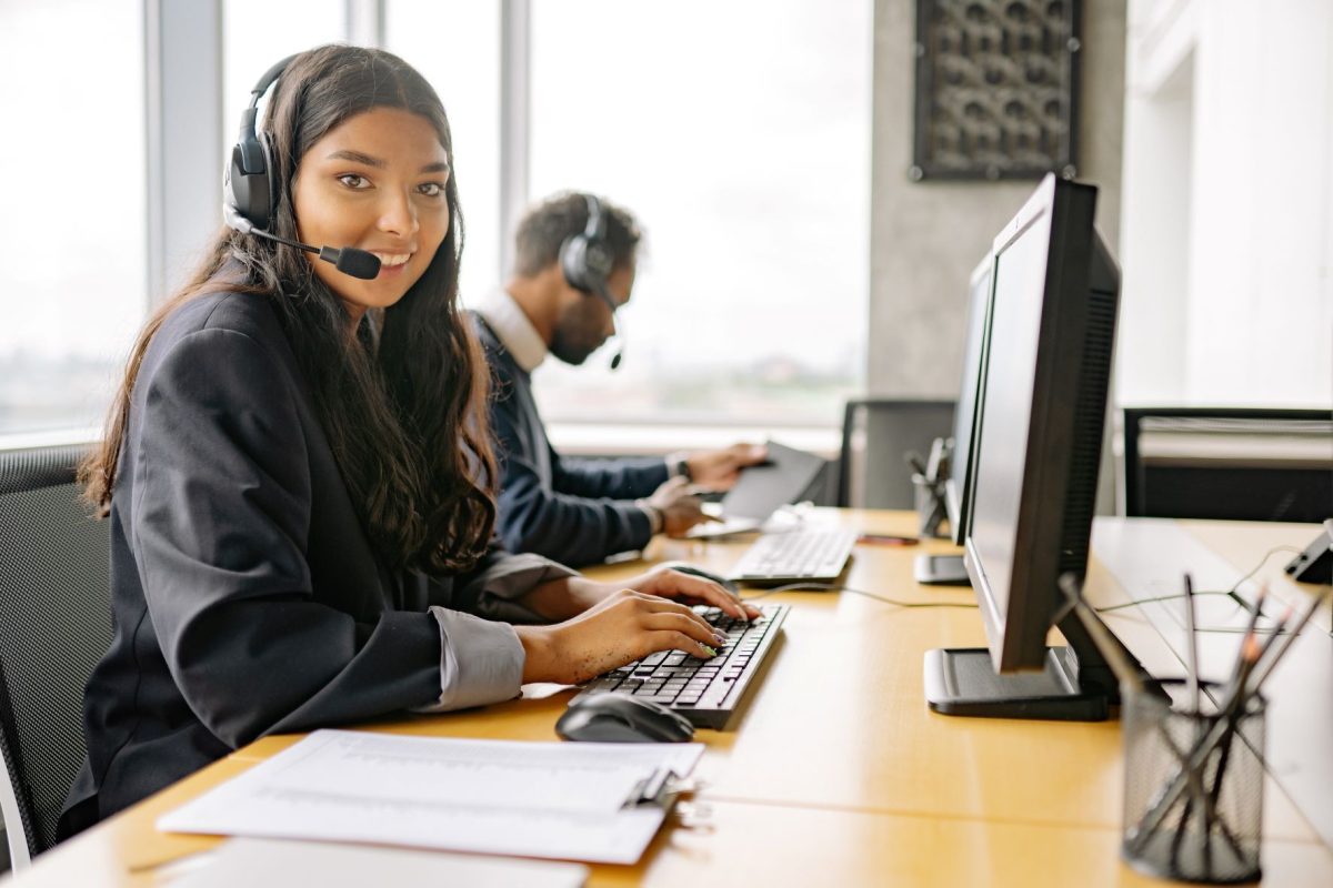 Smiling woman wearing a headset typing at a desk in a call center with a computer monitor nearby