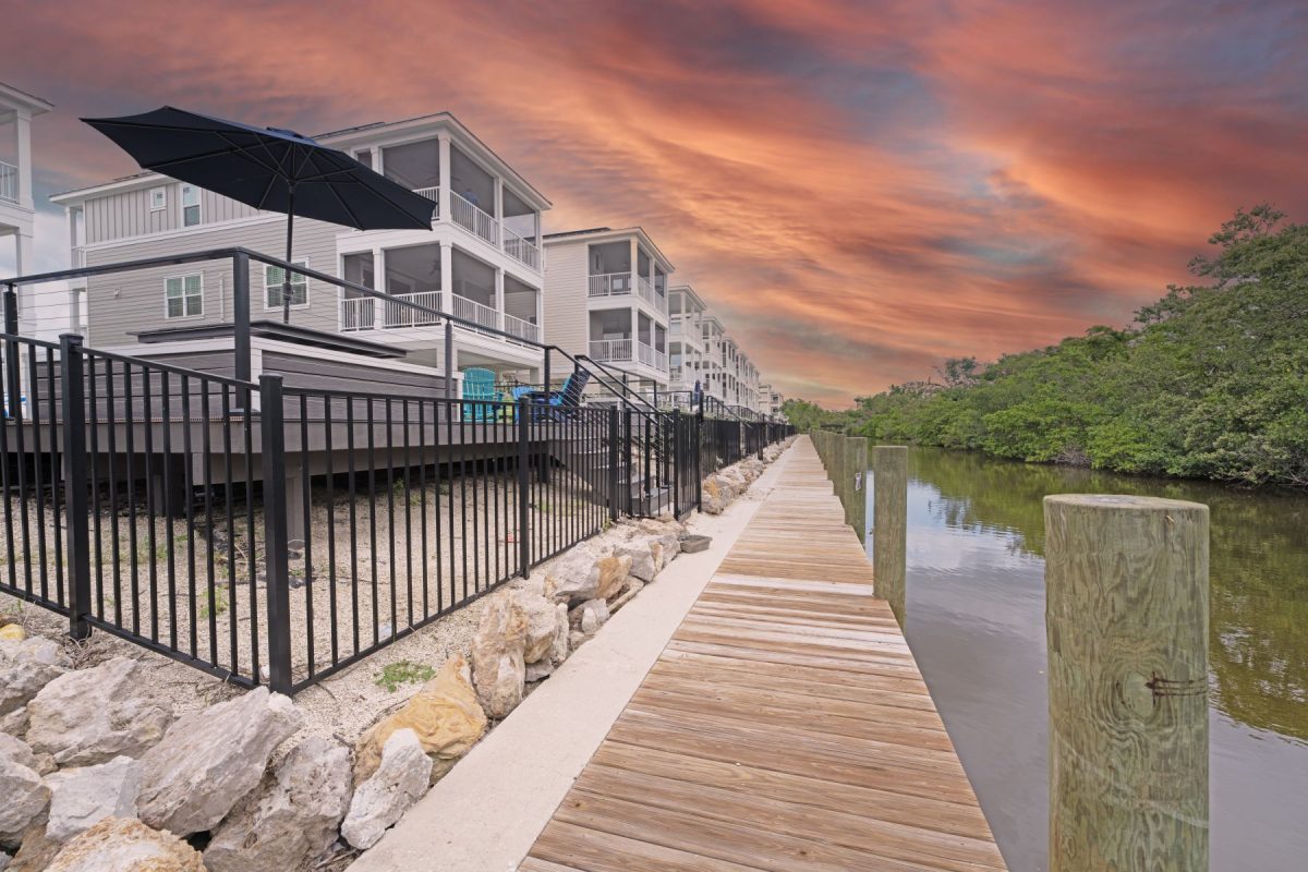 Boardwalk along a waterfront with modern gray houses, a black railing, and an orange sunset sky.